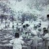 Foto arsip cetak hitam-putih jadul dan buram dari Majalah Api Kartini tahun 1962, memperlihatkan puluhan anak-anak usia dini sedang berkumpul dan membuat lingkaran besar sambil berpegangan tangan di bawah pepohonan rindang Lapangan Polonia dalam kegiatan Taman Minggu Melati, didampingi seorang pengasuh perempuan di sisi kanan halaman (ardiholmes.id).