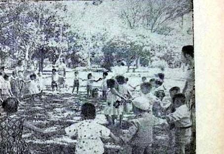 Foto arsip cetak hitam-putih jadul dan buram dari Majalah Api Kartini tahun 1962, memperlihatkan puluhan anak-anak usia dini sedang berkumpul dan membuat lingkaran besar sambil berpegangan tangan di bawah pepohonan rindang Lapangan Polonia dalam kegiatan Taman Minggu Melati, didampingi seorang pengasuh perempuan di sisi kanan halaman (ardiholmes.id).