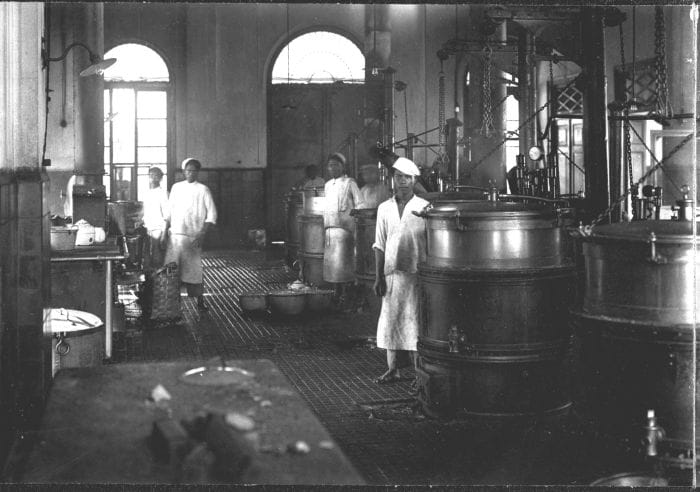 Foto hitam-putih interior dapur besar berteknologi uap di Krankzinnigengesticht Soember Porong (RSJ Lawang) Jawa Timur era kolonial Hindia Belanda periode 1902-1922. Tampak beberapa pekerja pribumi mengenakan pakaian dan penutup kepala putih berdiri di antara instalasi ketel uap besi raksasa untuk memasak makanan massal pasien.