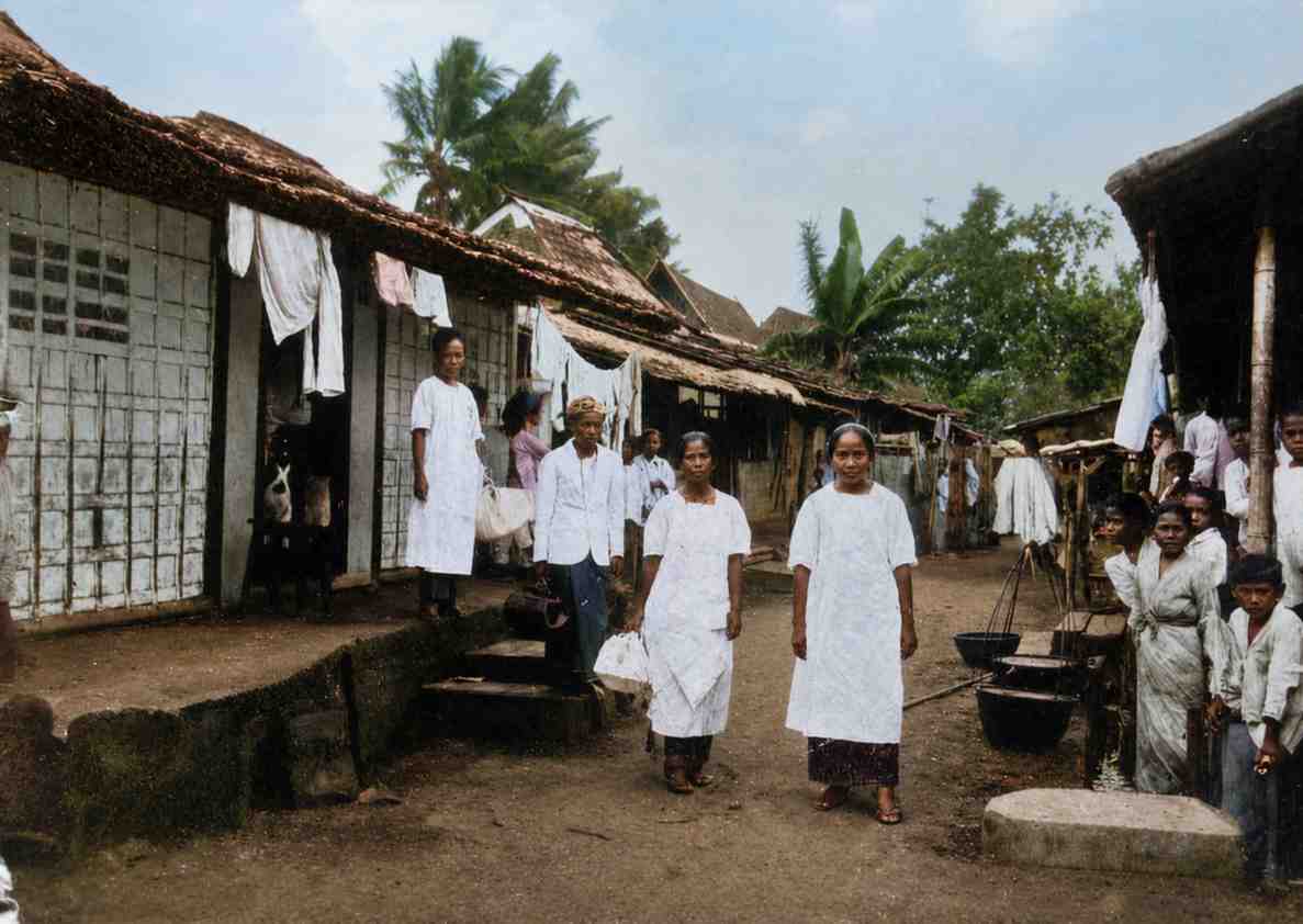 Foto berwarna (diedit) era kolonial menampilkan dua bidan bergaun putih bersih berjalan di gang sempit perkampungan rumah bambu Batavia yang dipenuhi warga lokal dan anak-anak (ardiholmes.id)