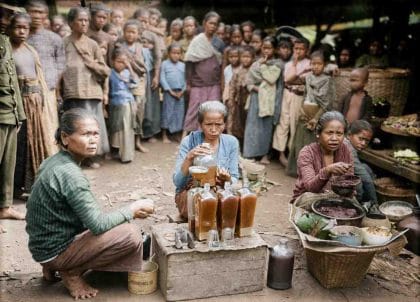 Foto kolase sejarah diwarnai yang memperlihatkan tiga perempuan lansia pribumi sedang duduk meracik jamu tradisional di ruang terbuka. Perempuan di tengah menuangkan cairan jamu kecokelatan ke dalam botol kaca besar menggunakan corong, sementara di sekeliling mereka puluhan warga desa dan anak-anak berkumpul menyaksikan. Di lantai tanah tampak wadah penumbuk dan bakul berisi tanaman herbal (ardiholmes.id).