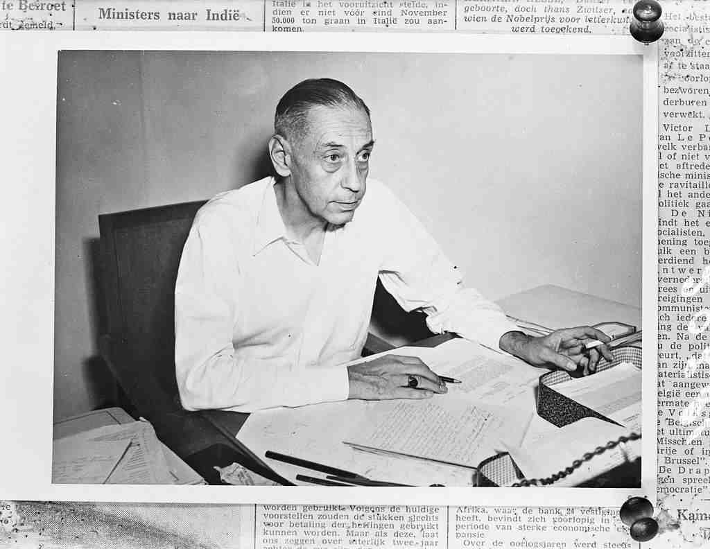 Foto hitam-putih Profesor P.M. van Wulfften Palthe mengenakan kemeja putih, sedang duduk di meja kerja menghadap dokumen dan memegang pulpen.