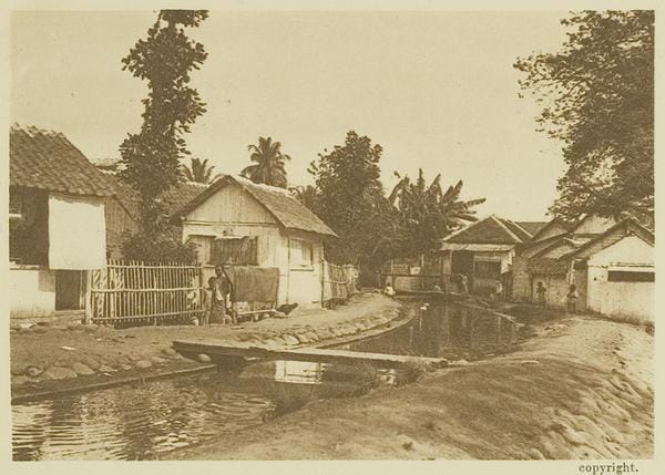 Foto kartu pos lama bernuansa sepia yang menggambarkan lanskap sebuah kampung di Malang sekitar tahun 1925. Tampak deretan rumah tinggal sederhana dari kayu dan bambu beratap rumbia di sepanjang aliran kanal air kecil, dengan latar belakang pepohonan tropis dan beberapa penduduk lokal yang berdiri di depan rumah.