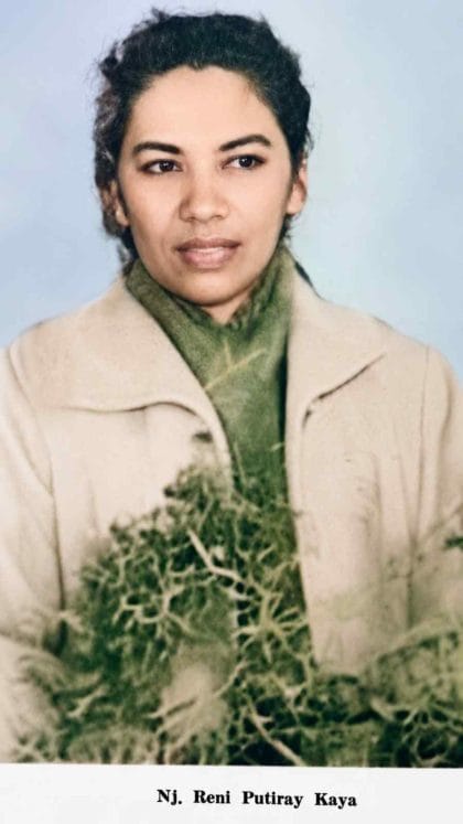 Potret setengah badan Nj. Reni Putiray Kaya, tokoh pergerakan perempuan dan komponis lagu anak-anak asal Maluku. Beliau mengenakan mantel musim dingin berwarna krem, syal tebal hijau, dan memegang seikat dedaunan dengan latar belakang biru muda cerah. Terdapat teks cetak nama di bagian bawah foto. (ardiholmes.id)