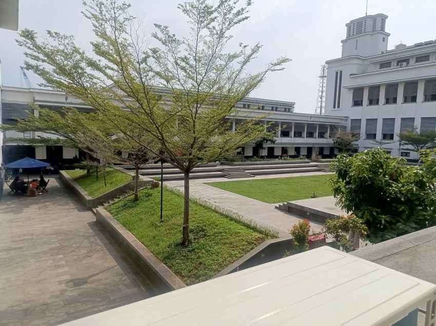 Halaman dalam Museum Bank Indonesia berupa taman rumput hijau dengan pohon rindang dan pagar tanaman, dikelilingi sayap gedung putih berlantai dua serta menara tandon bergaya art deco menjulang di sudut kanan kompleks; beberapa pengunjung duduk di bawah payung biru (ardiholmes.id).