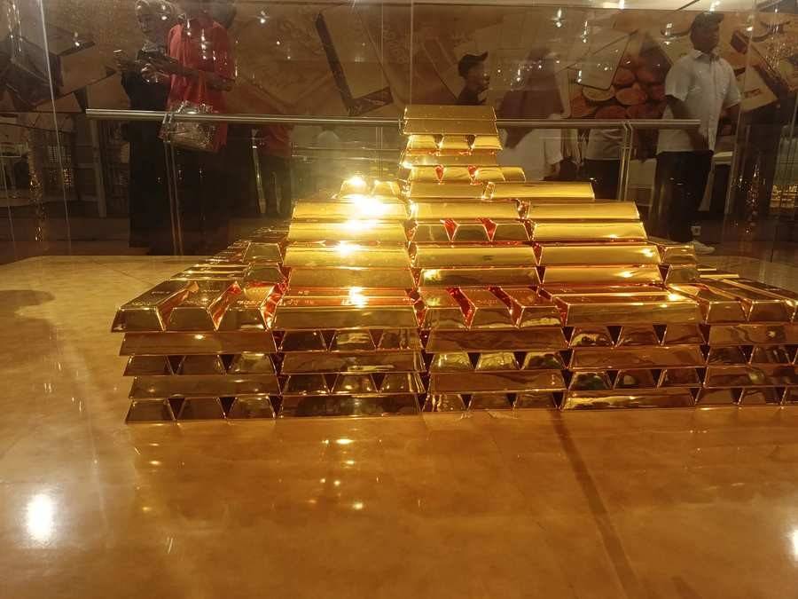 Tumpukan replika batangan emas tersusun membentuk piramida di dalam ruang kaca Museum Bank Indonesia, memantulkan cahaya keemasan ke lantai, dengan beberapa pengunjung tampak samar terpantul di dinding kaca sedang memotret (ardiholmes.id).
