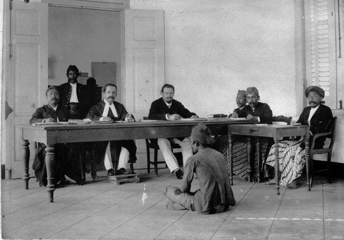 Foto arsip sebuah sidang Landraad di Hindia Belanda sekitar 1890-1900. Landraad adalah pengadilan bagi penduduk pribumi. Tampak hakim ketua Eropa duduk di balik meja panjang bersama anggota pengadilan pribumi, dengan hadirin duduk di ruangan terbuka. ardiholmes.id